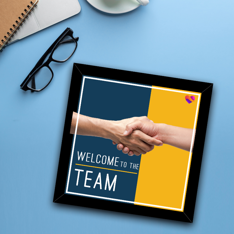 Framed poster with handshake graphic and 'Welcome to the Team' text on a blue surface with glasses and notebook.