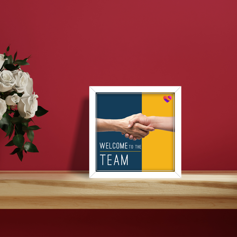 Framed poster with handshake graphic and 'Welcome to the Team' text on a red wall with flowers.
