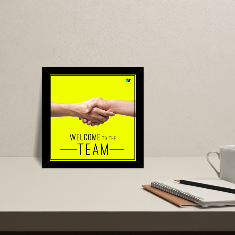 Framed poster with handshake graphic and 'Welcome to the Team' text on a yellow background, placed on a desk.