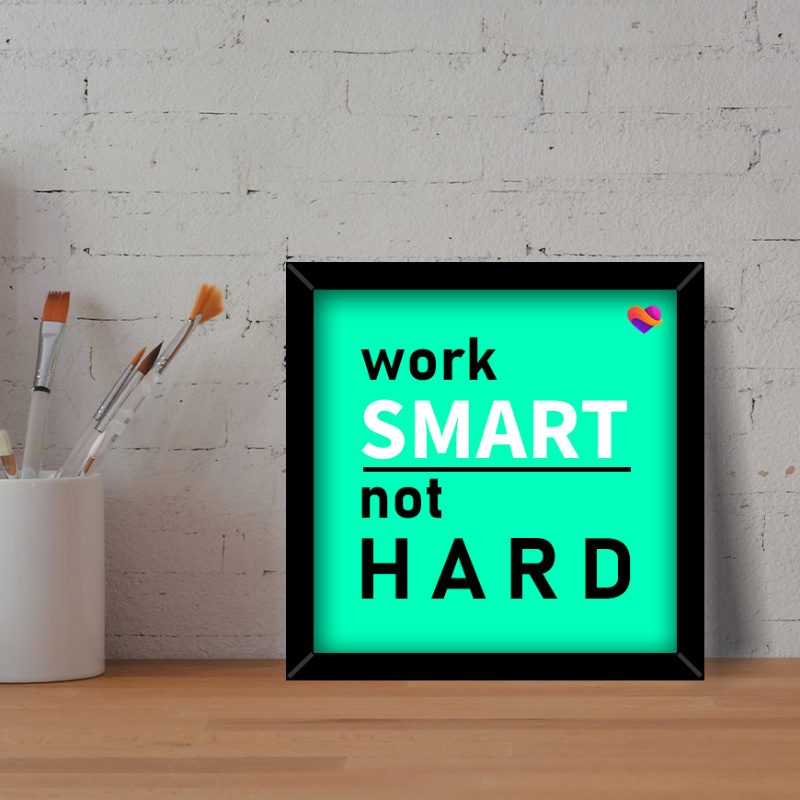 Framed poster with 'work SMART not HARD' text on a light green background, placed on a wooden surface with a white container and brushes.