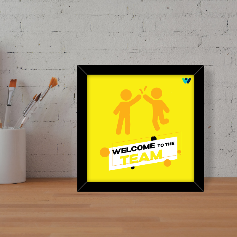 Framed poster with 'Welcome to the Team' text and two people giving a high-five on a yellow background, placed on a wooden surface with a white brick wall in the background.