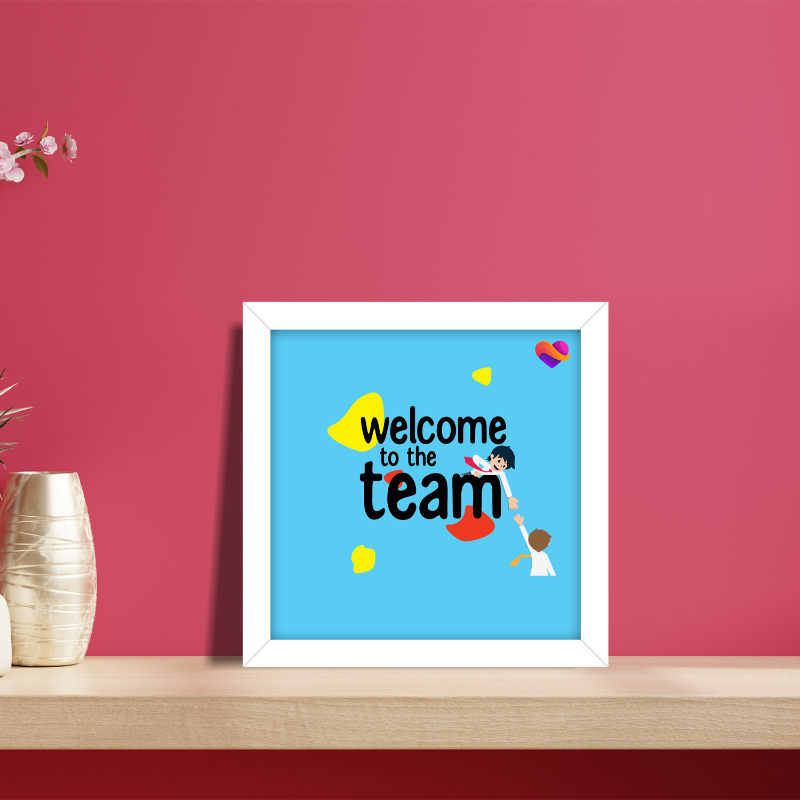 Framed artwork with 'Welcome to the team' text on a blue background, placed on a wooden surface against a red wall.
