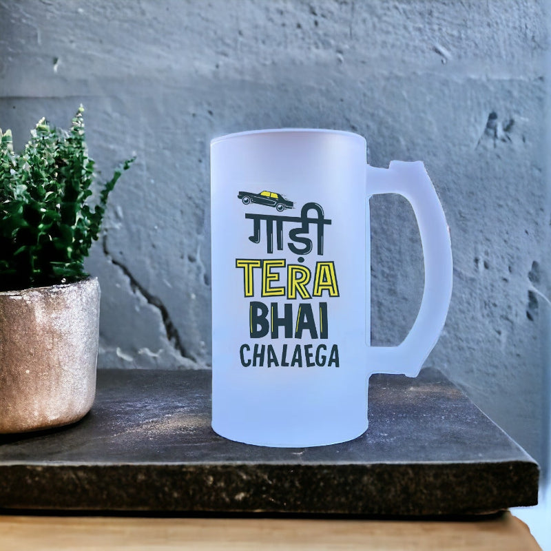 A white ceramic beer mug with the text 'Gaadi Tera Bhai Chalayega' printed in green and yellow colors, placed on a table with a plant and a concrete surface in the background.