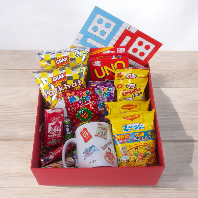 A red gift hamper basket containing various snack foods and a Ludo game piece, all arranged on a wooden surface.