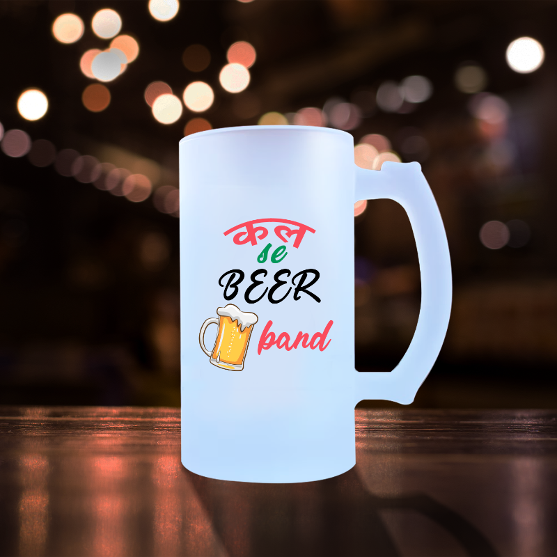 A white ceramic beer mug with the text 'Kal se BEER Band' and a graphic of a beer glass, set against a blurred background with Christmas lights.