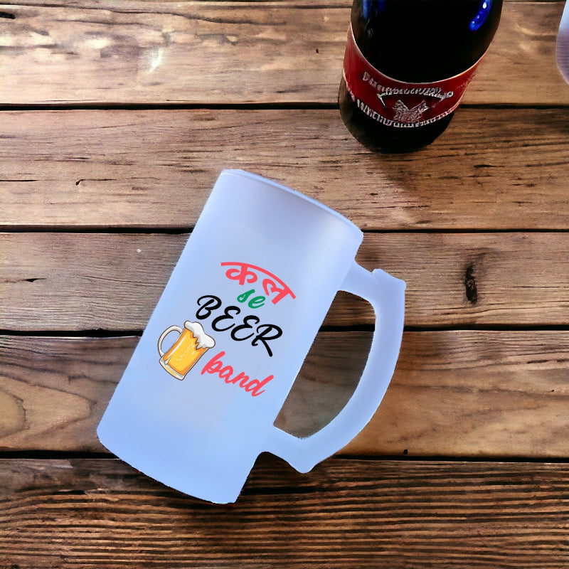 Frosted mug with colorful text and a beer graphic on a wooden surface next to a bottle.