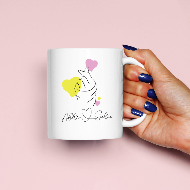A person holding a white ceramic mug with a heart and 'Abhi' printed on it, with navy blue nails and a pink background.
