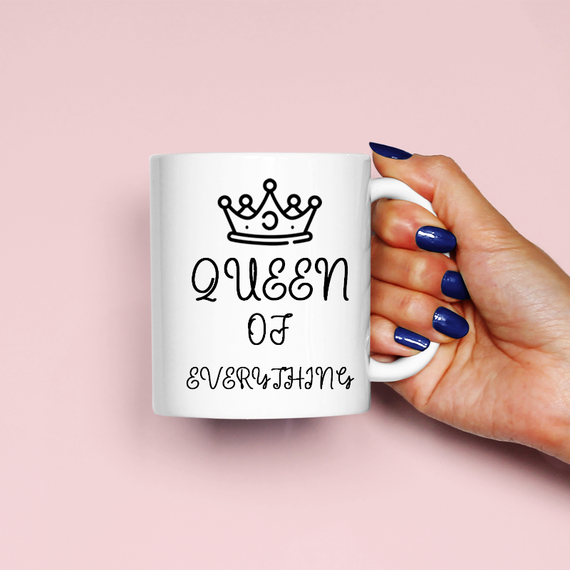 White mug with 'Queen of Everything' text and a crown, held by a hand with blue nail polish on a pink background.