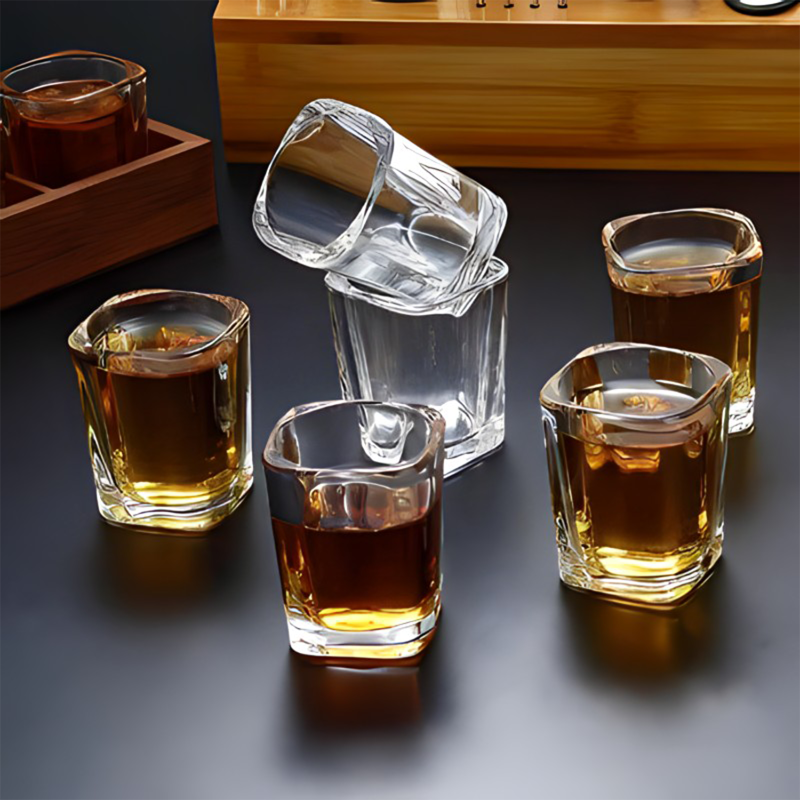 A set of six clear glass shot glasses with a heavy base, displayed on a dark surface. The glasses contain a liquid, possibly a shot of alcohol, and are positioned in a line.