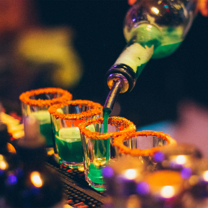 Green liquid being poured into shot glasses with orange rims on a bar.