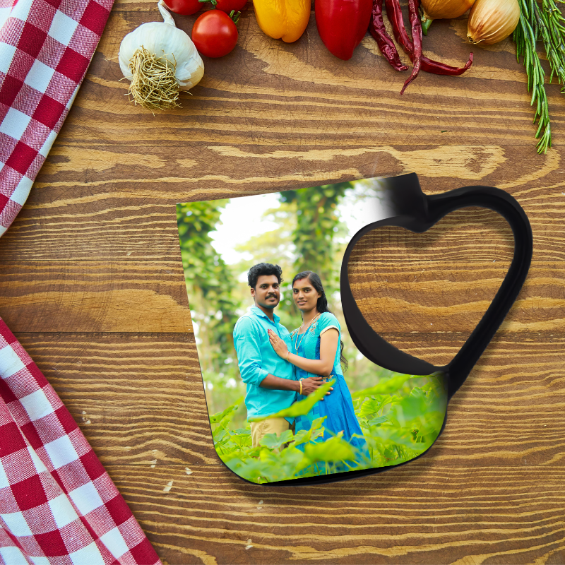 A heart-shaped ceramic mug with a couple's photo on it, placed on a wooden surface with a checkered cloth in the background, surrounded by various vegetables.