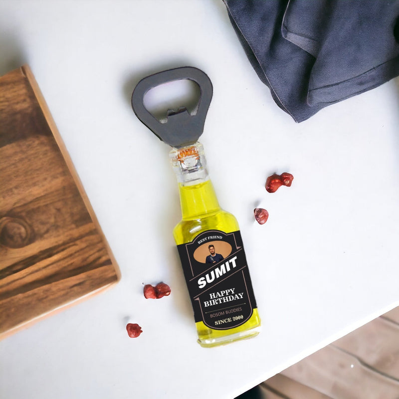 A personalized bottle opener with a happy birthday message, a bottle with a yellow liquid, and a metal bottle opener on a table with some red berries scattered around.