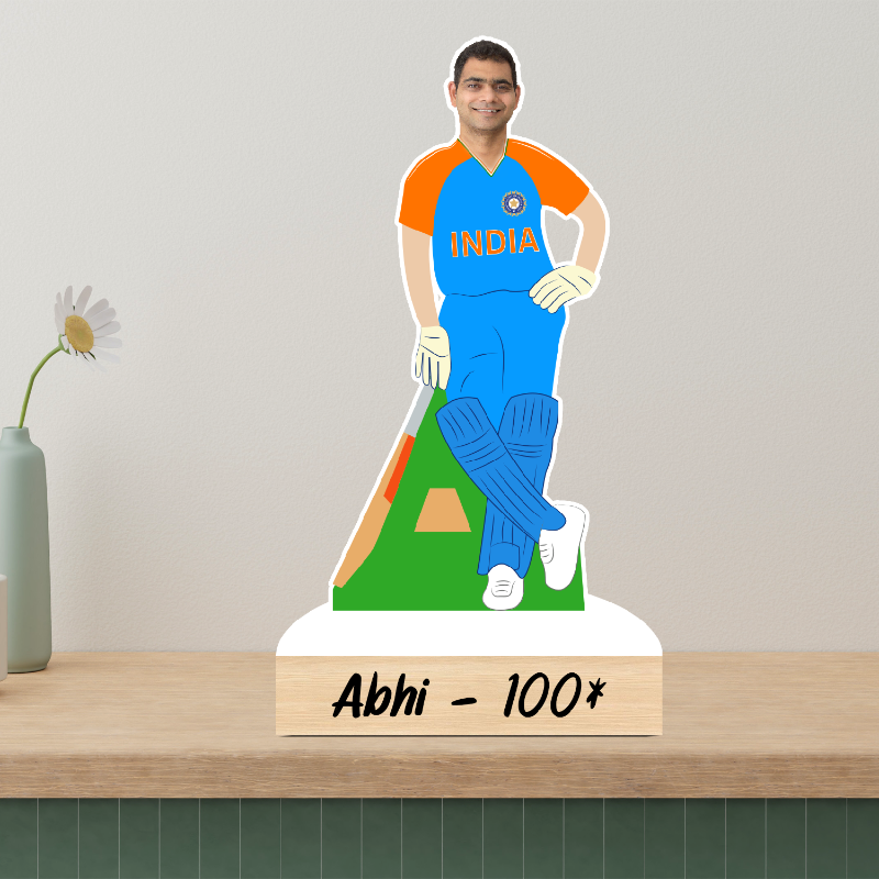 Ceramic figurine of a cricket player in India jersey on a wooden shelf with a vase and flower in the background.