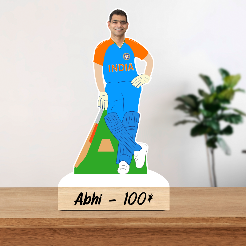 Cricketer figure in India jersey on a stand with 'Abhi - 100*' text, against a white wall with a plant.