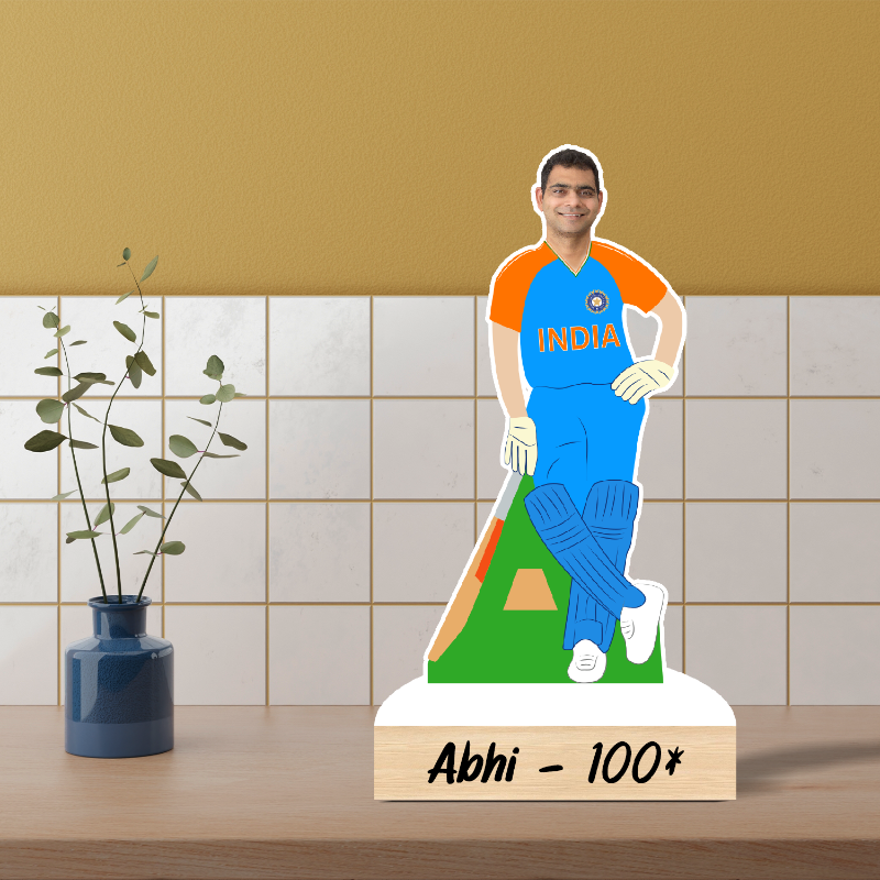 Ceramic figure of a cricketer in an India jersey on a wooden stand with 'Abhi - 100*' text, against a tiled wall background.