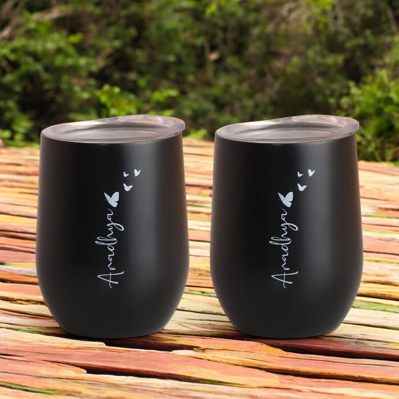Two black insulated mugs with custom names printed on them, placed on a wooden surface with greenery in the background.