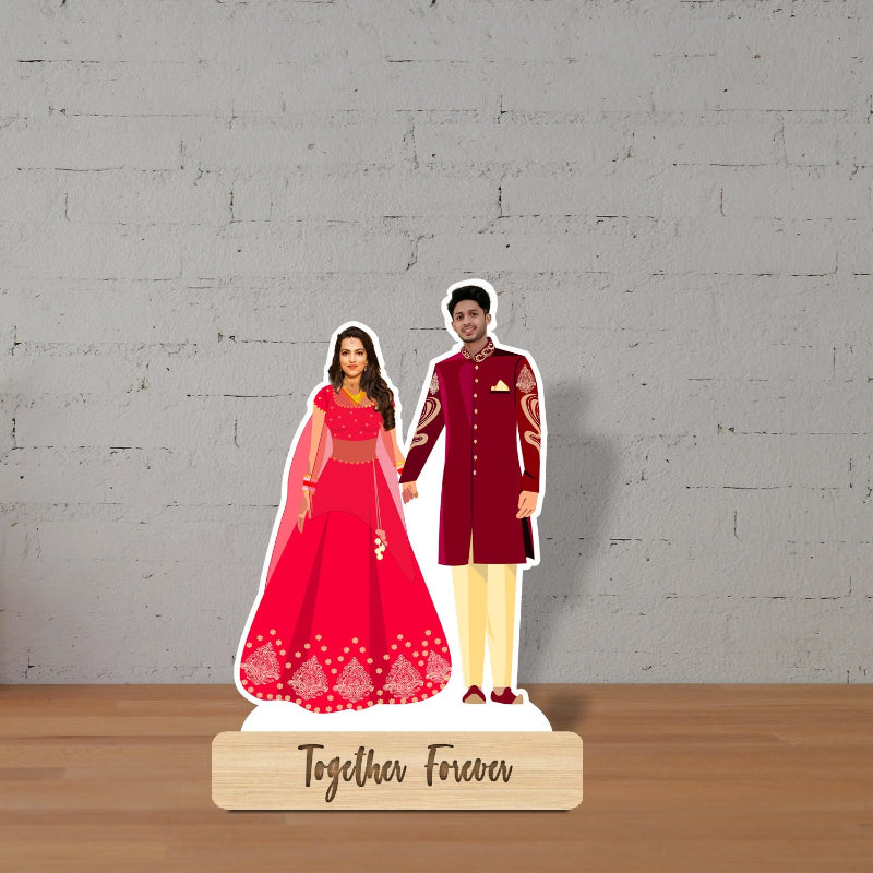 Cardboard cutout of a couple in traditional attire with 'Together Forever' text on a wooden stand against a gray brick wall.