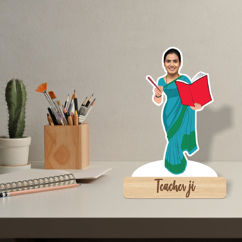 Desk setup with a paper cutout of a teacher holding a book, a pen holder, and a cactus plant.