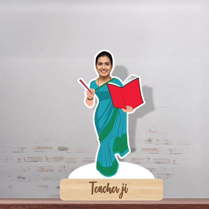 A personalised caricature of a lady teacher standing on a wooden stand with a red book, wearing a green and blue sari. The caricature is smiling and has a book titled 'Teacher Ji' on the wooden base.