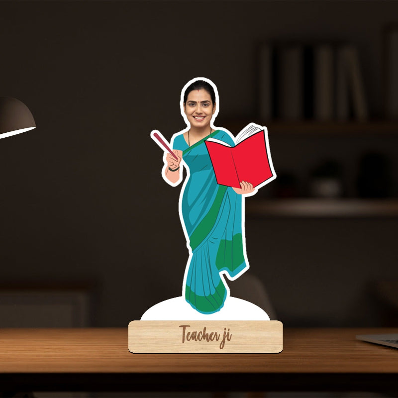 Decorative figure of a woman in a blue saree holding a red book and a pointer, on a wooden base with 'Teacher ji' text, against a blurred indoor background.