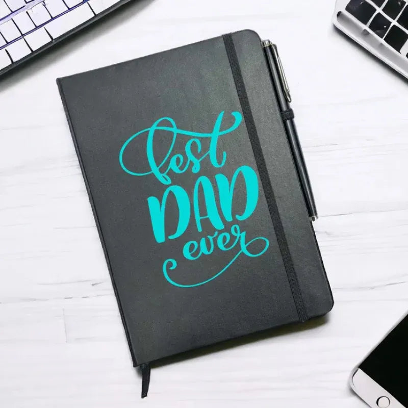 Black notebook with 'Best Dad Ever' in teal on a white surface with computer equipment.