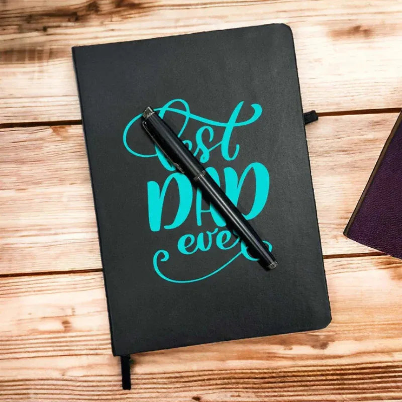 Black notebook with 'Best Dad Ever' text and a pen on a wooden surface