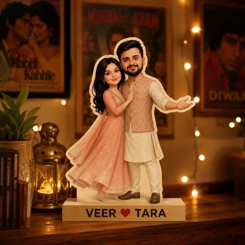 Personalized couple figurine of a man and woman standing together on a wooden base with the text 'VEER ♥ TARA'. The man is dressed in a beige kurta and vest, and the woman is in a flowing pink dress.