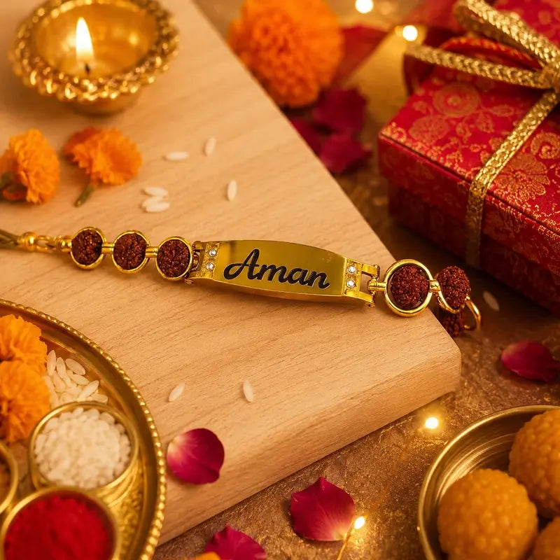 A gold-plated personalized Rakhi bracelet with the name 'Aman' engraved in black script, adorned with red and purple textured beads, displayed on a wooden board surrounded by festive decorations including marigolds, a lit diya, and a red gift box.