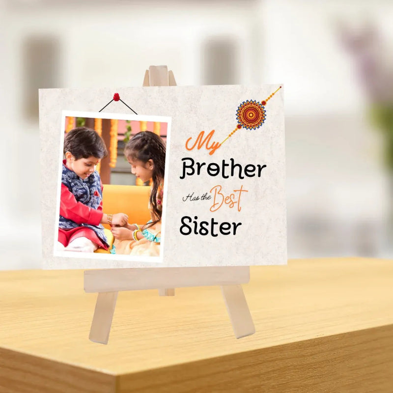 A personalized photo print displayed on a small wooden easel. The print features a placeholder photo of a boy and a girl, alongside the text 'My Brother Has the Best Sister' in black and orange lettering, with a decorative orange and red Rakhin (wheel of fortune) illustration in the top right corner.