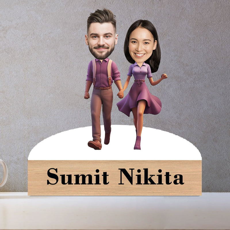 Personalized bobblehead figures of a man and woman holding hands with a wooden nameplate below on a gray background.
