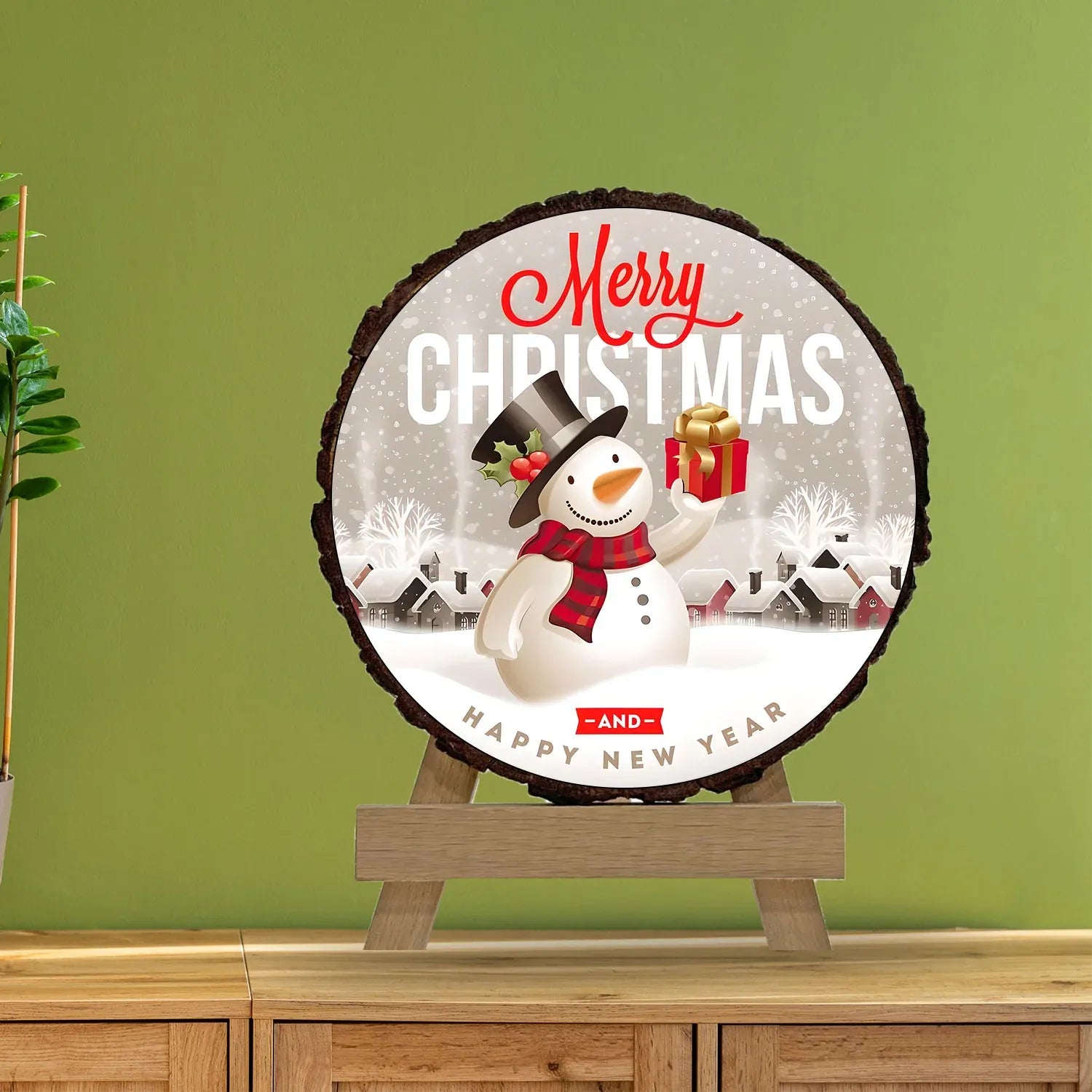 Round Christmas-themed sign with a snowman on a wooden stand against a green wall.