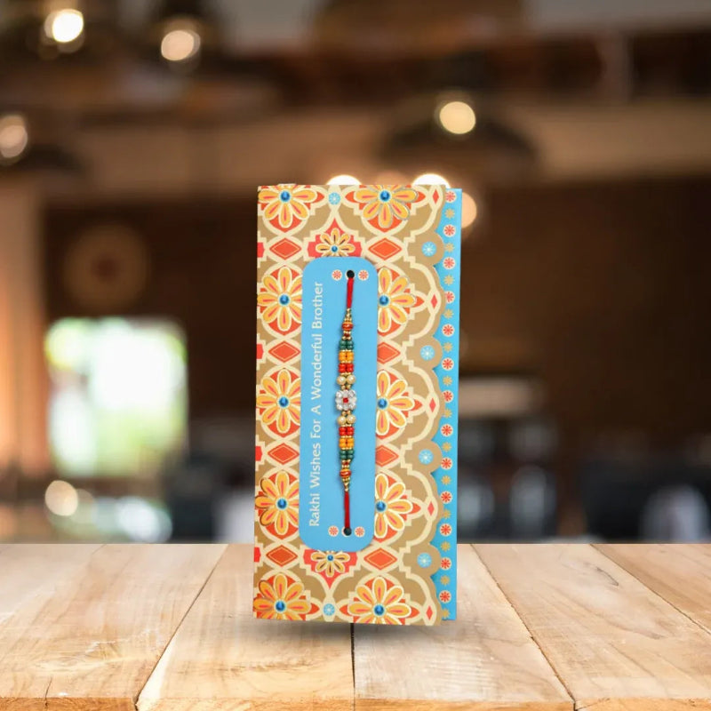 A colorful patterned gift box standing on a wooden table, with a small beaded Rakhi thread visible through a blue rectangular window. The box has an ornate floral and geometric design in orange, beige, blue, and red, with the text 'Rakhi Wishes For A Wonderful Brother' printed vertically.