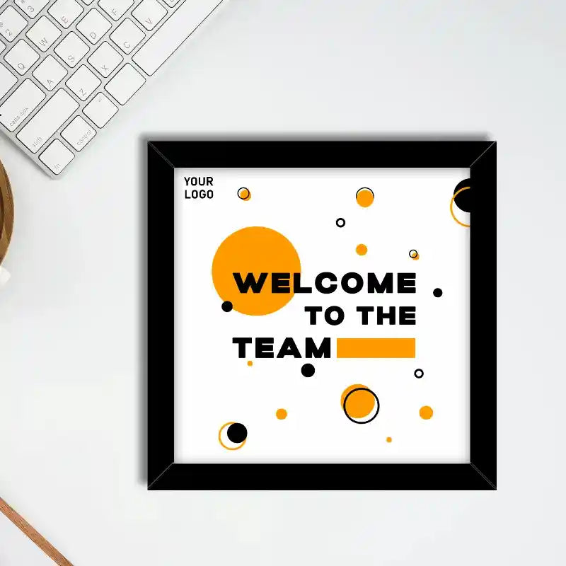 A corporate photo frame: framed square wall art print with the text 'WELCOME TO THE TEAM' in black, set on a white desk next to a keyboard. The print features a white background with abstract orange and black circles and is enclosed in a black frame.