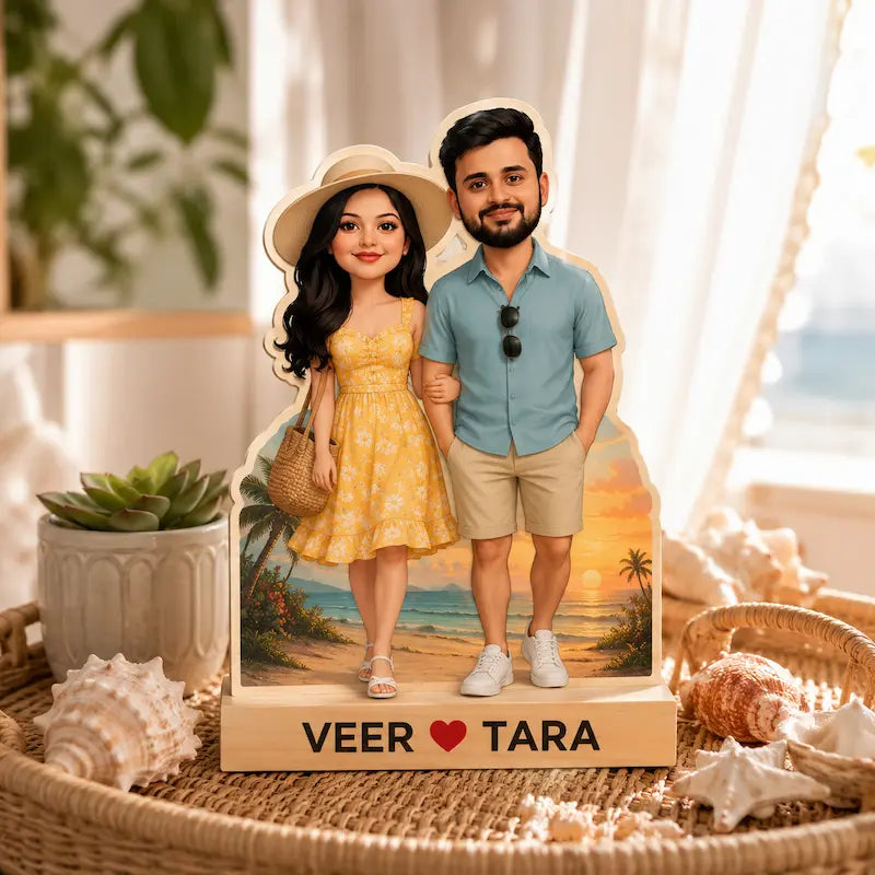 Customizable 2D cartoon couple figurines standing on a personalized wooden base with the names 'VEER' and 'TARA' and a heart symbol. The couple is dressed in a yellow dress and a blue shirt with khaki shorts, set against a tropical beach backdrop.