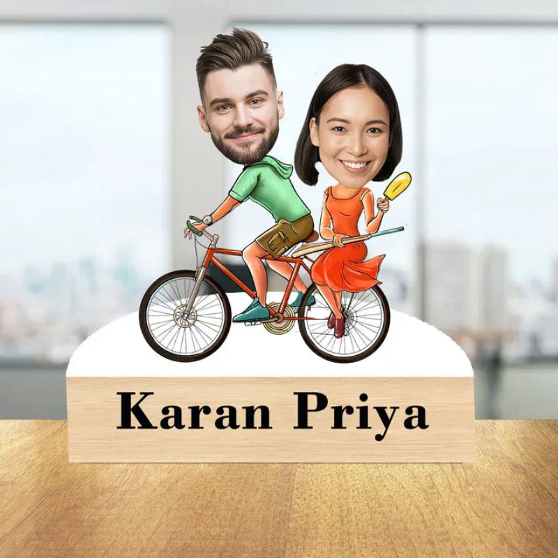 Caricature of a man and woman on a bicycle with 'Karan Priya' below, set against a blurred indoor background.