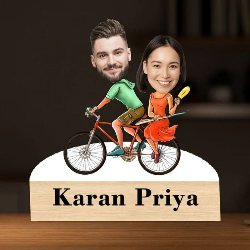 Bobblehead figures of a man and woman on a bicycle with 'Karan Priya' below, against a dark background.