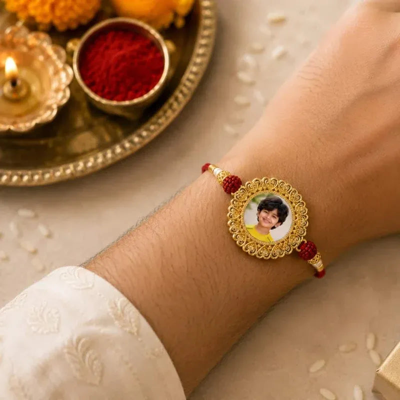 A close-up of a person's wrist wearing a gold and red traditional Indian rakhi bracelet with a custom photo charm, set against a festive background with a brass plate, lit diya, and red powder.