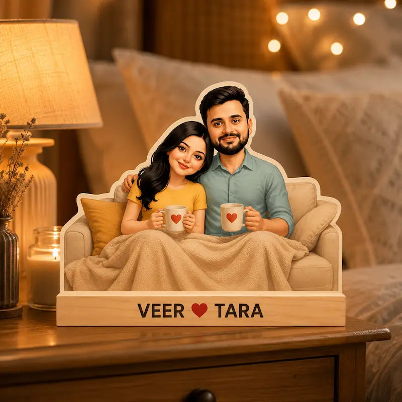A custom cardboard cutout couple's sign showing a cartoon-style man and woman sitting on a sofa under a blanket, each holding a mug with a heart, mounted on a wooden base printed with 'VEER ♥ TARA'. The sign is styled on a wooden table in a cozy, warmly lit bedroom setting.