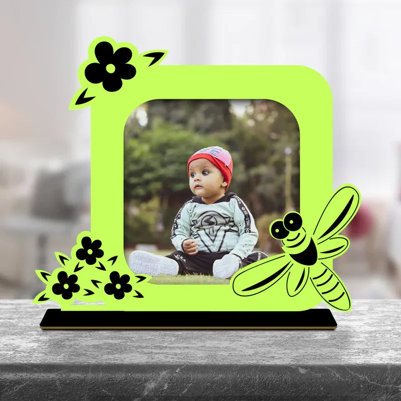 Cute baby photo frame gift — neon green acrylic standee with bee and floral cartoon design, custom baby photo printed inside square opening, black base, indoor setting.