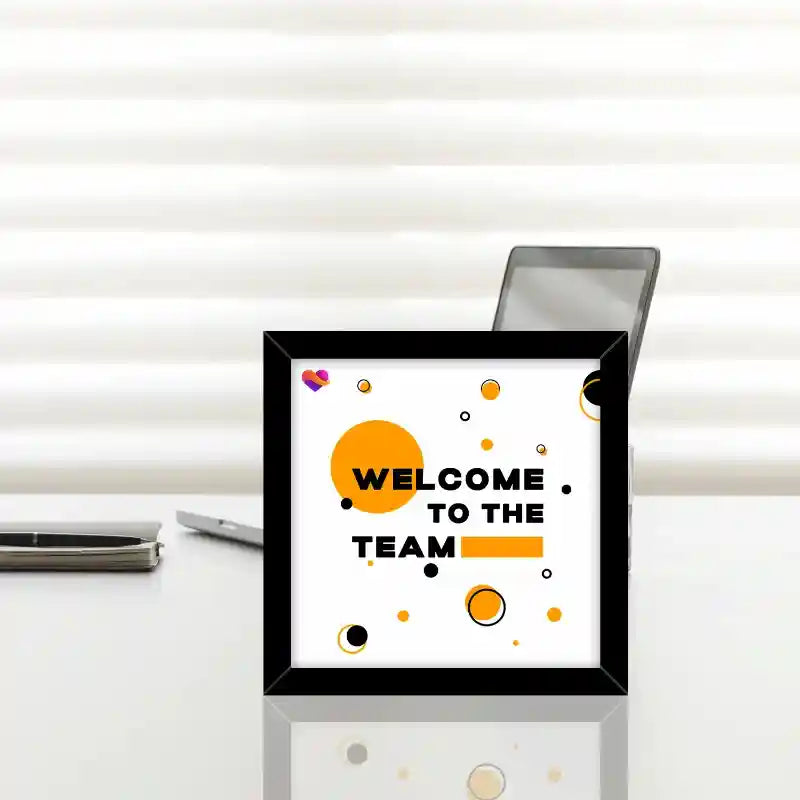 A framed square wall art print with the text 'WELCOME TO THE TEAM' in black on a white background, decorated with orange and black circles, displayed on a white desk with a laptop and phone behind it.
