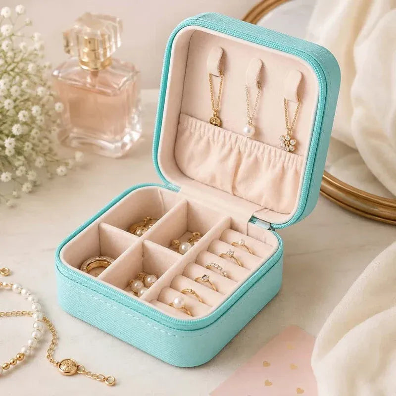 A turquoise zippered jewelry box with a cream interior, shown open and filled with rings, earrings, and pearl-like jewelry. The box is photographed on a light tabletop with soft decorative props, including a perfume bottle, flowers, and draped fabric.