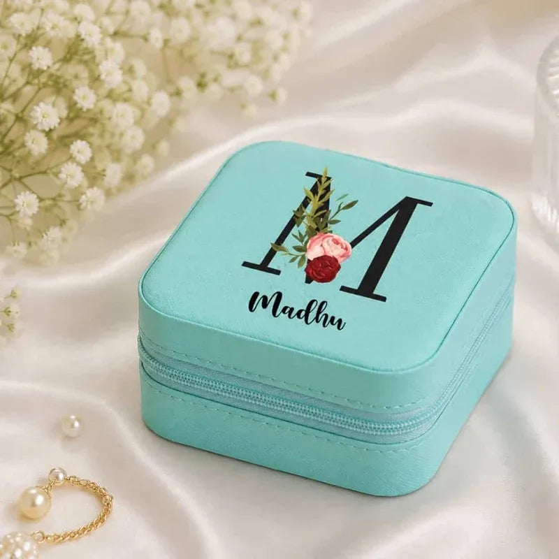 A square turquoise jewelry box with a zipper closure, decorated with a large black monogram letter 'M', the name 'Madhu' in script, and a small floral design. The box is styled on a white satin fabric background with baby's breath flowers and a pearl necklace nearby.