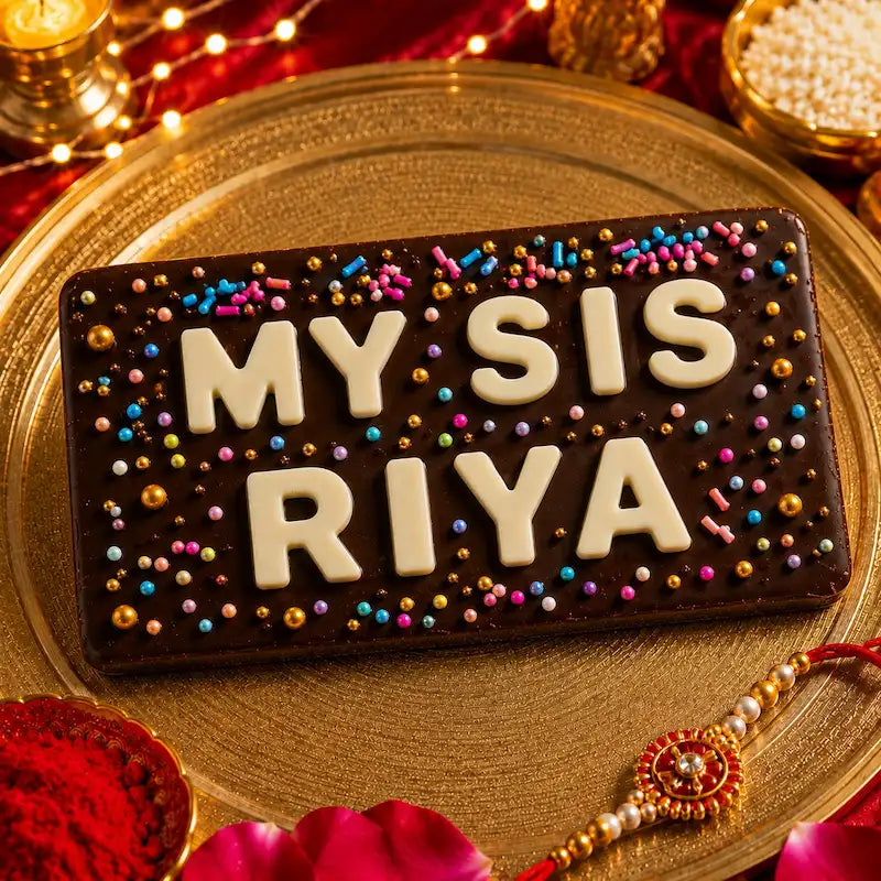 A rectangular chocolate cake or plaque with white lettering reading 'MY SIS RIYA' and decorated with colorful sprinkles, presented on a gold platter with festive decorations including a Rakhi, powder bowl, and string lights.