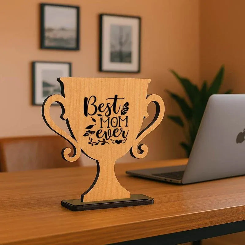 Wooden trophy with 'Best Mom Ever' engraving on a desk with a laptop and framed pictures in the background.