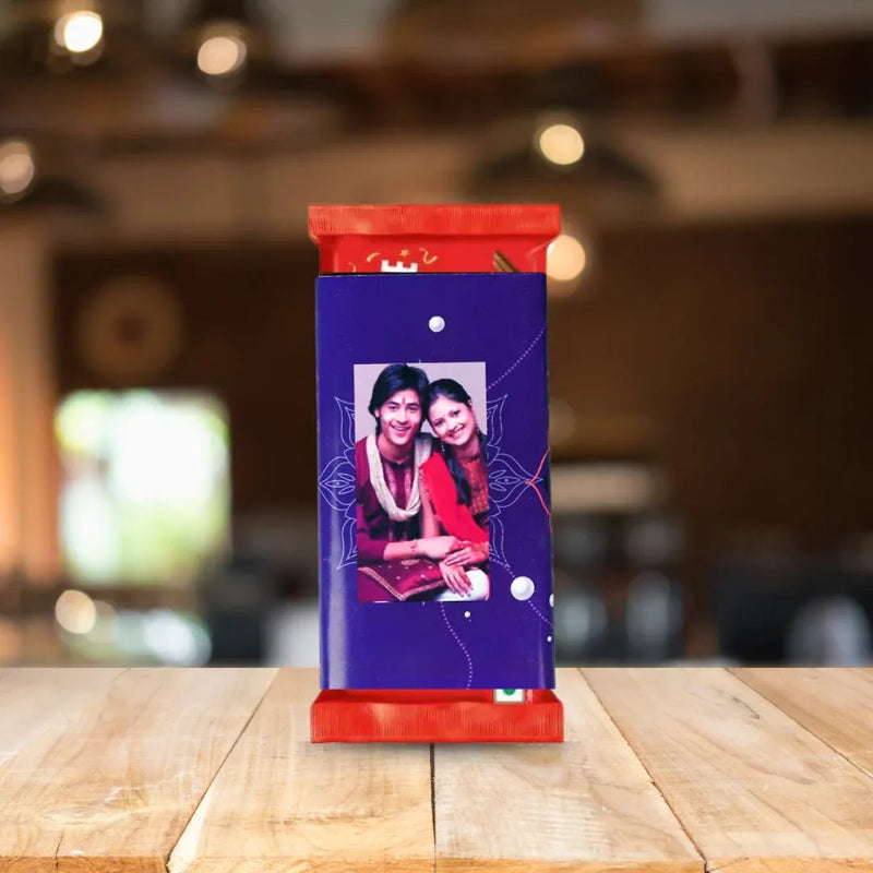 A custom photo chocolate bar package standing upright on a wooden table. The wrapper is primarily deep purple with a printed photo of two women in red tops, framed by a red top and bottom border, with subtle white decorative line details.