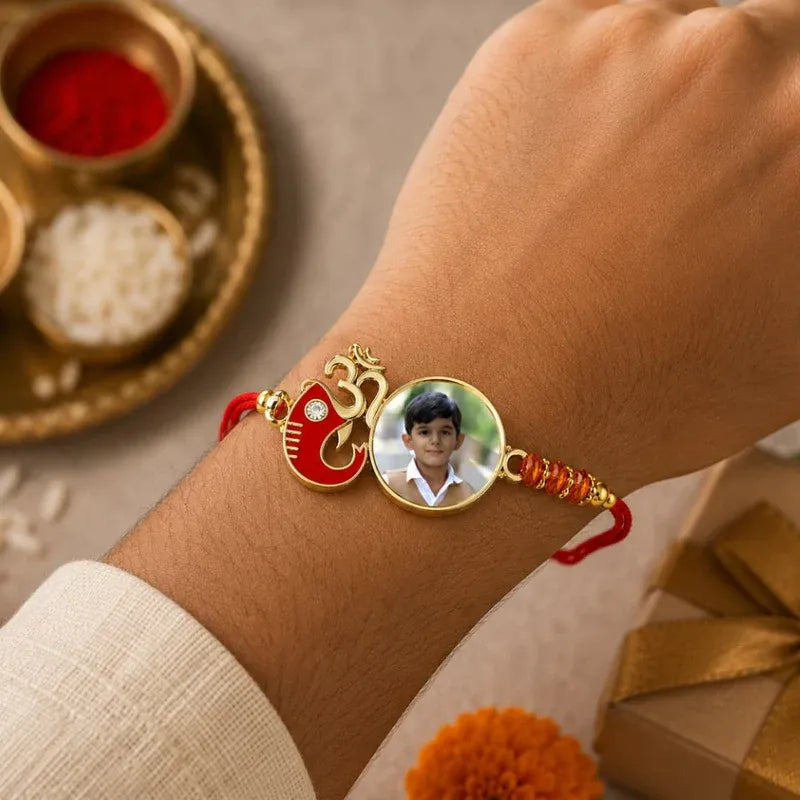 A custom photo frame Rakhi bracelet worn on a person's wrist, featuring a red and gold Om symbol charm and a circular photo insert, with traditional festive props in the background.