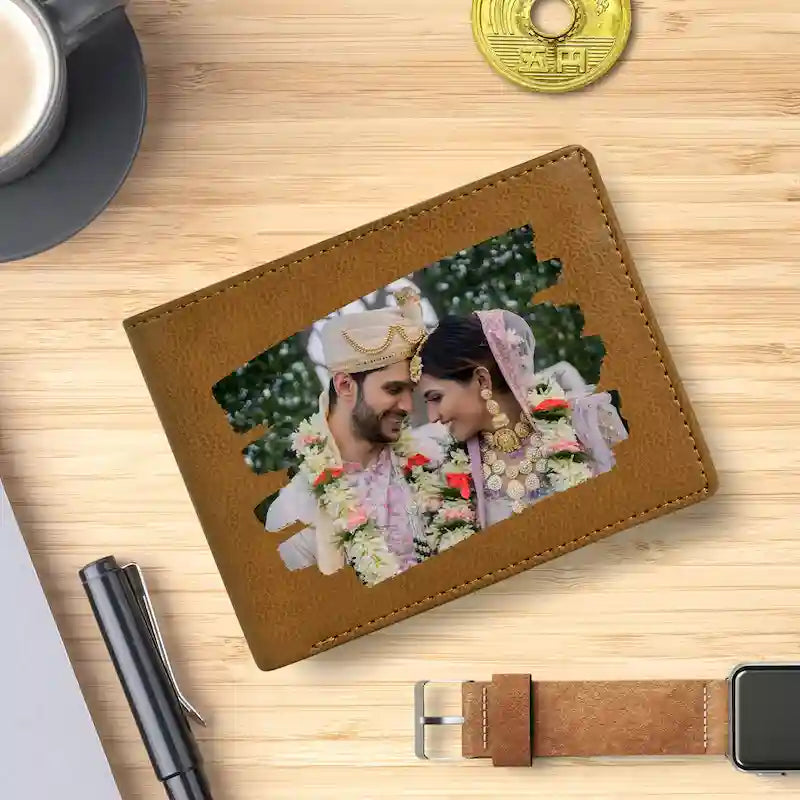A brown custom photo wallet for men or card holder lying on a light wood desk, printed with a custom photo of a bride and groom. Surrounding props include a pen, a smartwatch with a matching brown leather band, a coin, and a cup on a saucer.