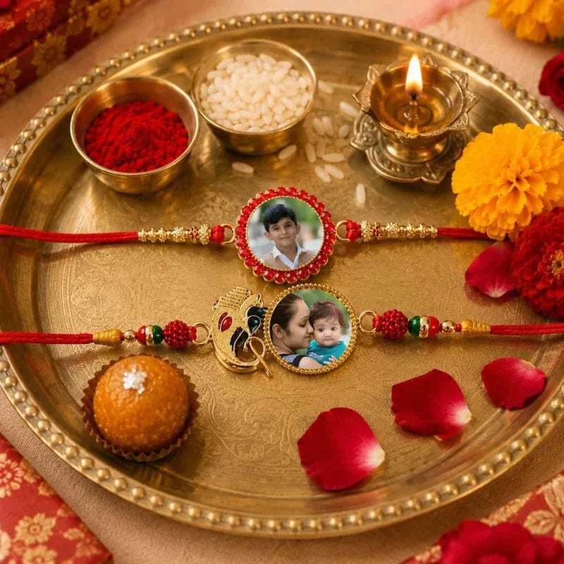 Two personalized Rakhi wristlets with photo frames are displayed on a traditional brass tray with red powder, rice, a burning diya, marigold flowers, and a laddoo.