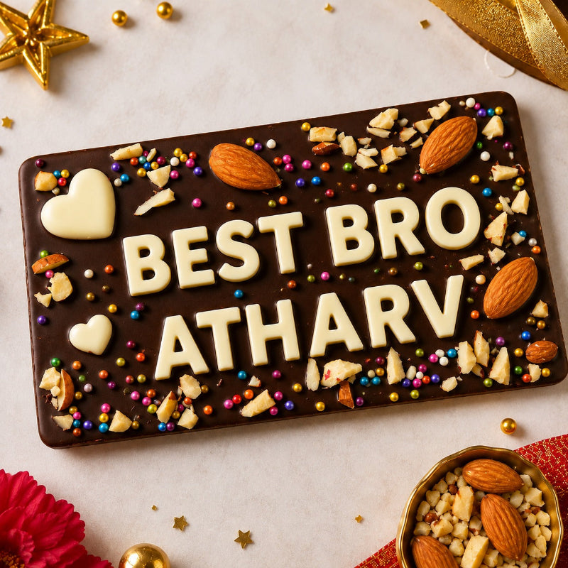 A rectangular chocolate bar with the text 'BEST BRO ATHARV' in white lettering, decorated with nuts, chocolate hearts, and gold foil flakes, set on a light background with festive props.