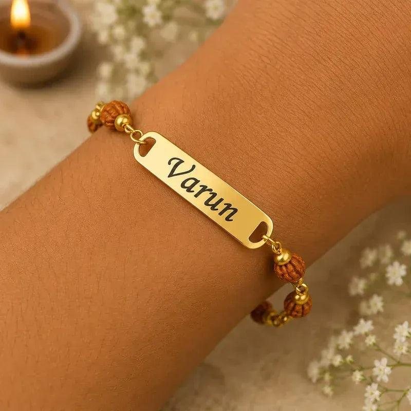 A gold-plated nameplate bracelet engraved with the name 'Varun', worn on a person's wrist. The bracelet features brown beaded accents on either side, set against a soft, warm background with blurred white flowers and a burning candle.
